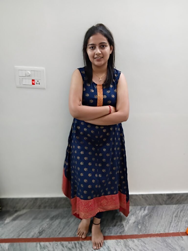 A young woman standing indoors against a plain white wall, smiling with her arms crossed. She is wearing a navy blue traditional kurta with golden patterns and a red border, along with dark leggings. She is barefoot and standing on a grey marble floor.